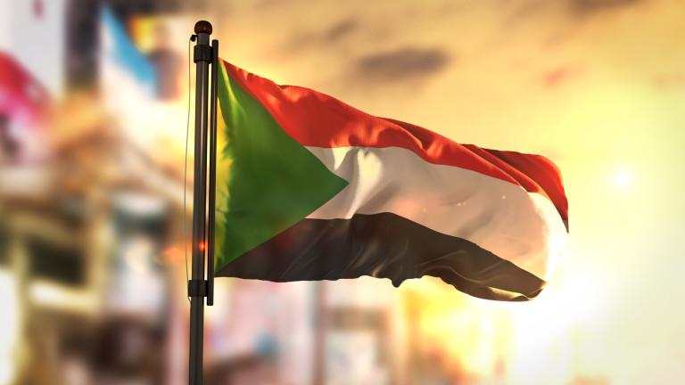 Sudan Flag Against City Blurred Background At Sunrise Backlight