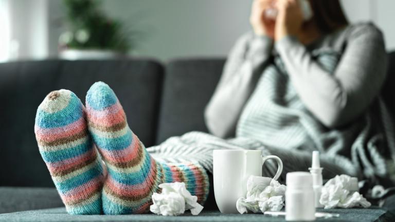 Sick woman with flu, cold, fever and cough sitting on couch at home. Ill person blowing nose and sneezing with tissue and handkerchief. Woolen socks and medicine. Infection in winter.